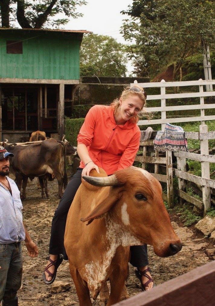 The author, Ketti Wilhelm, smiling, sitting on a cow in Nicaragua (while working as an international trip guide there), with a local cowboy standing by. ©KettiWilhelm2020