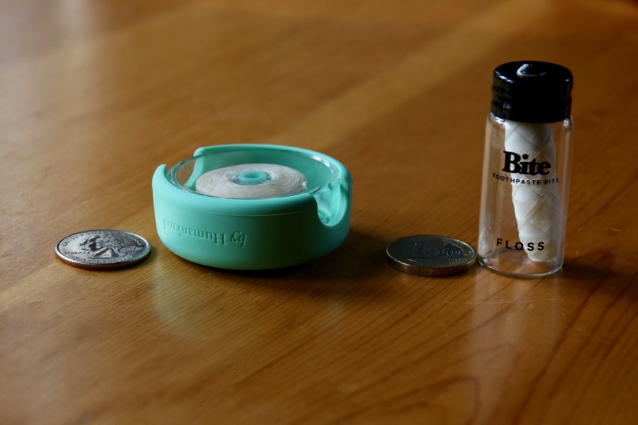 Two plastic-free dental floss options – byHumankind in a green, silicone and glass reusable container, and Bite, in a refillable glass jar – on a wooden table next to a quarter and a Euro for size comparison. ©KettiWilhelm2020