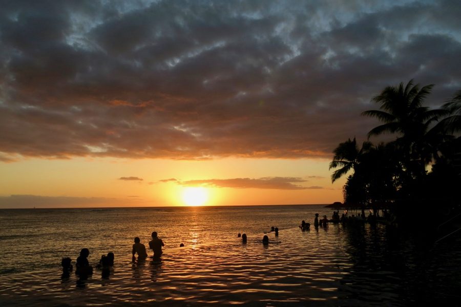 An orange sunset from an infinity pool on Oahu, Hawaii. Two people snap photos by the edge of the pool – it's clearly from pre-COVID travel days. ©KettiWilhelm2020