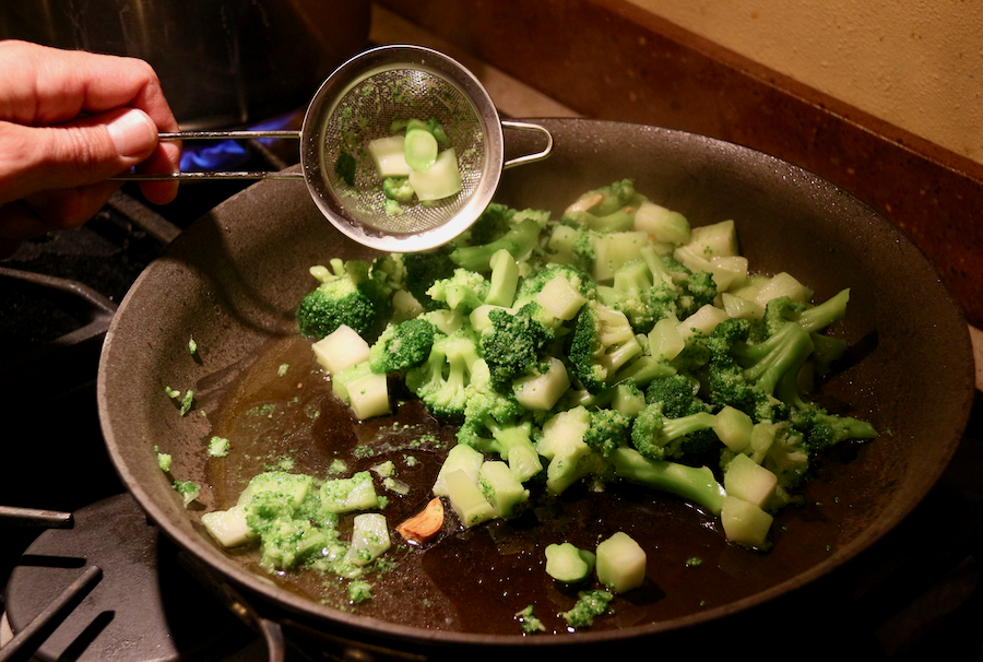 The boiled broccoli pieces being scooped out of the water and placed in the pan with the soffritto using a small colander. ©KettiWilhelm2021