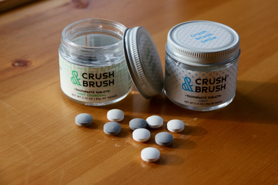 Glass jars of Crush & Brush Mint Charcoal and Mint toothpaste tablets, on a wooden table in the sun, reviewed here, from Nelson Naturals. ©KettiWilhelm2021