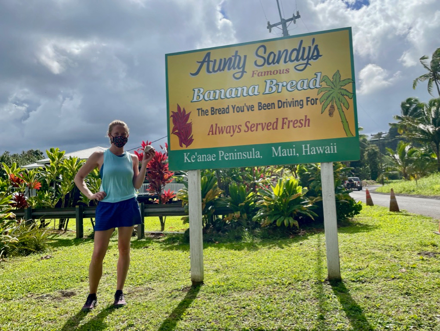 The author on Maui, wearing a mask, next to a sign for Aunty Sandy's banana bread, under stormy skies. ©KettiWIlhelm2021