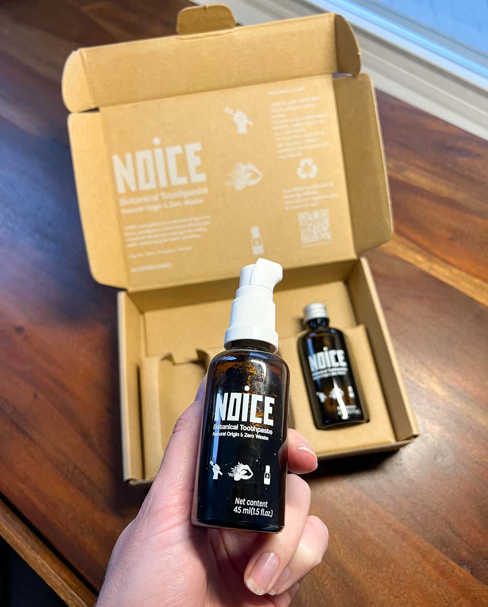 The blogger and review writer holds a glass jar of Noice organic, refillable toothpaste gel in hand, above the cardboard packing it came in, along with two plastic-free refill bottles. ©KettiWilhelm2022