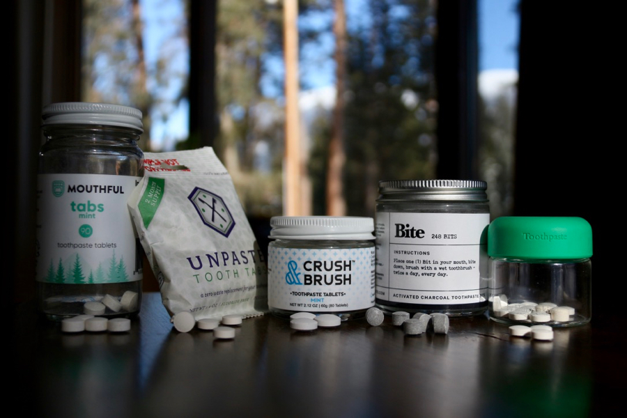 Glass jars and paper packets of plastic-free toothpaste tablets (Bite, Unpaste, Mouthful, Crush & Brush, and byHumankind brands) sitting on a stool with trees in the background. ©KettiWilhelm2021