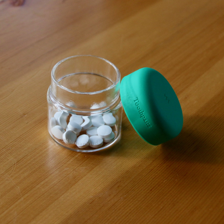 The glass and silicone refillable container for byHumankind's fluoride toothpaste tablets sits on a wooden table. ©KettiWilhelm2021