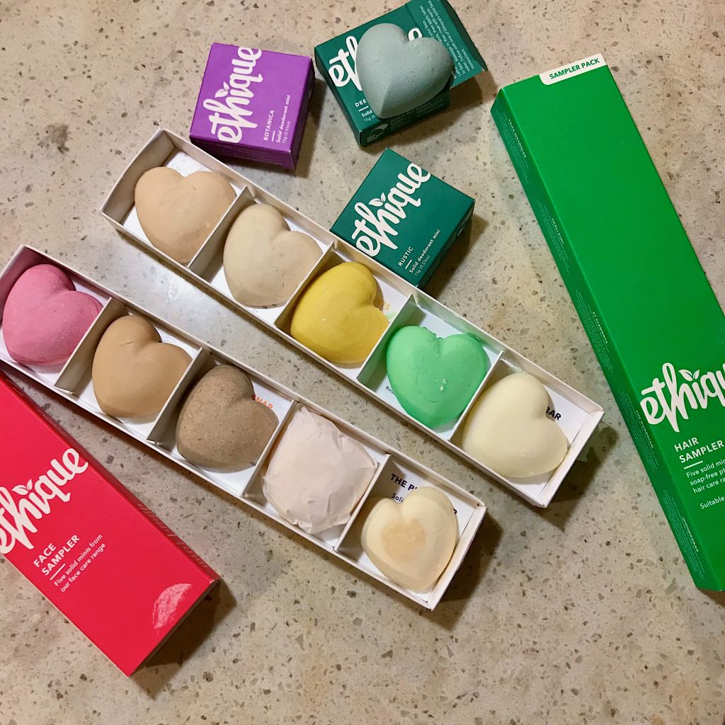 Ethique sample products – eco-friendly deodorant, shampoo bars, and solid face wash bars, all heart-shaped bars in paper packaging, laid out on a counter top. ©KettiWilhelm2021