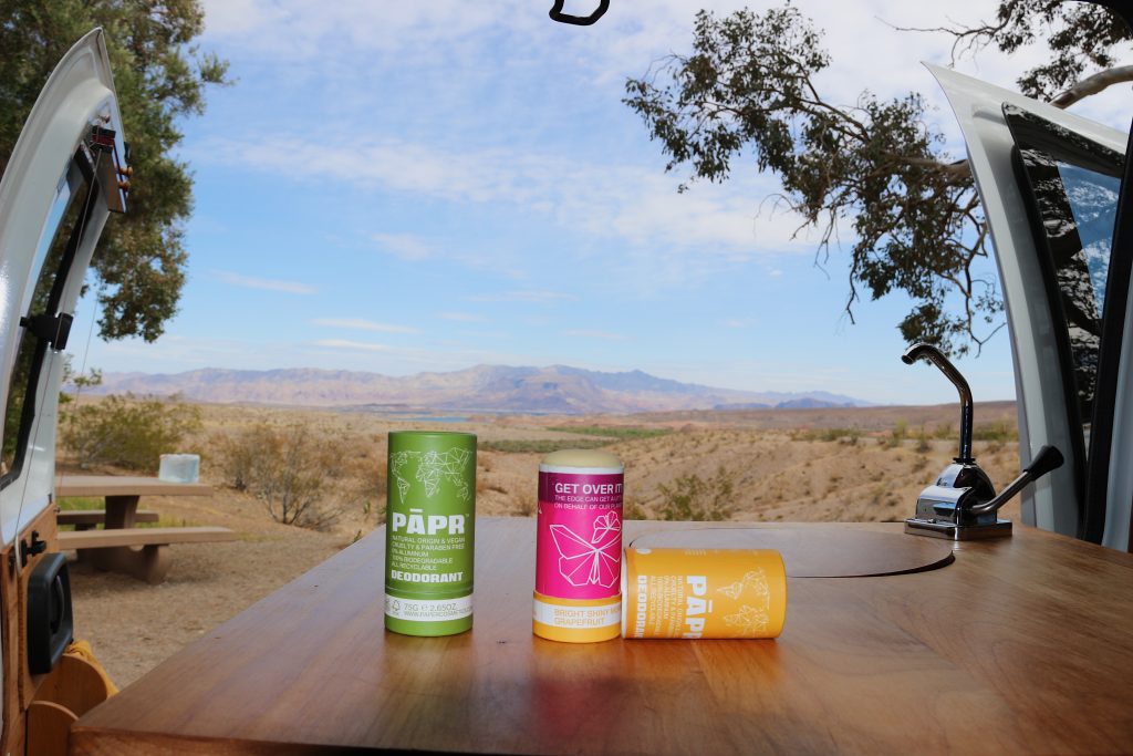 Two tubes of PAPR deodorant (plastic-free, in cardboard packaging) on a wood table with a sink inside a camper van, with the Nevada desert behind them. ©KettiWilhelm2021