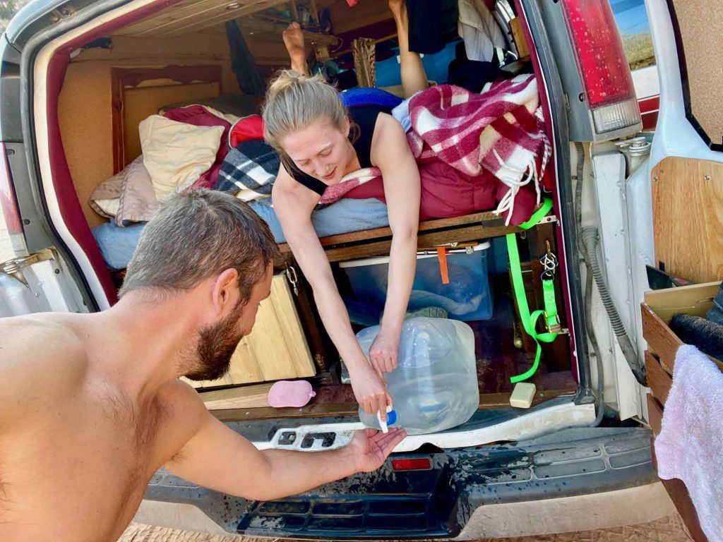 The author lying down in the bed of a homemade camper van, helping her husband open the spigot on a large plastic water bottle to a take an outdoor shower. ©KettiWilhelm2021