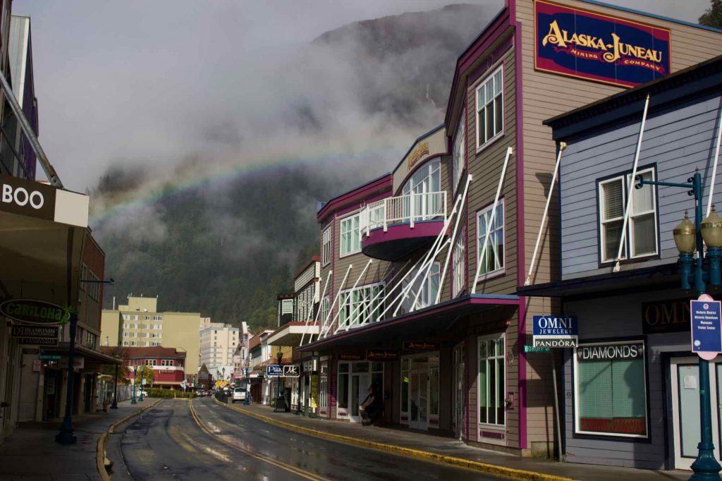 A rainbow forms in downtown Juneau, over a narrow, curved street filled with touristic shops. ©KettiWilhelm2021