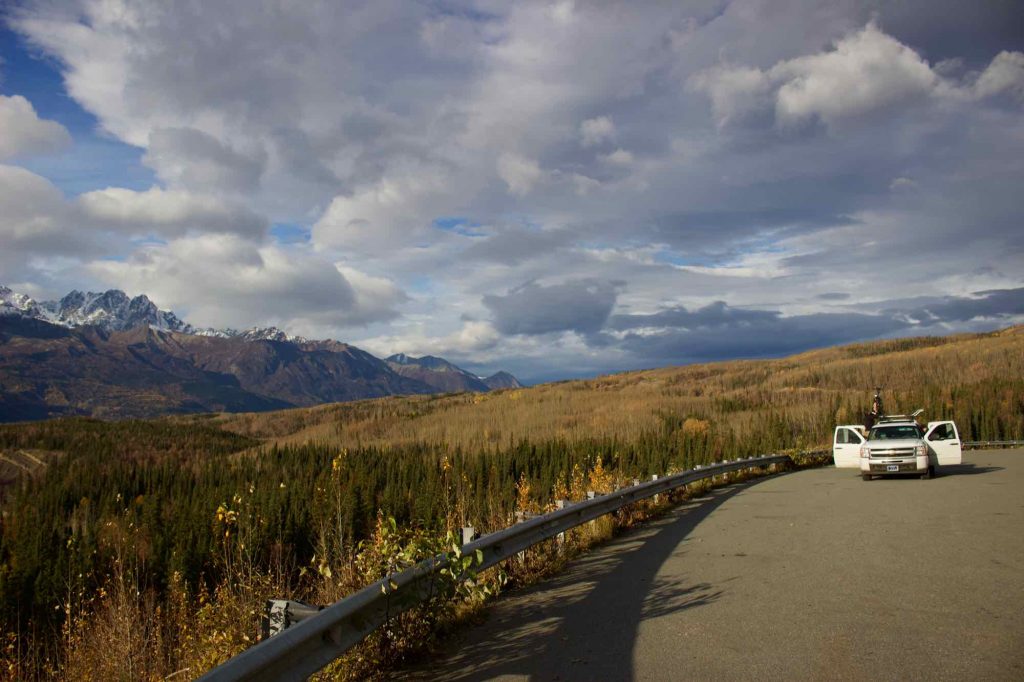 The white pickup truck that the author used for a road trip around Alaska, parked by a guardrail, next to an expansive view of a wide river valley and mountains. ©KettiWilhelm2021