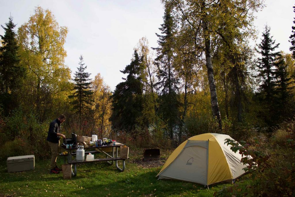 On the right, the yellow and white tent that the blogger slept in while traveling in Alaska, and her husband cooking dinner at a camp stove on a picnic table on the left, with yellow-leafed autumn trees behind. ©KettiWilhelm2021