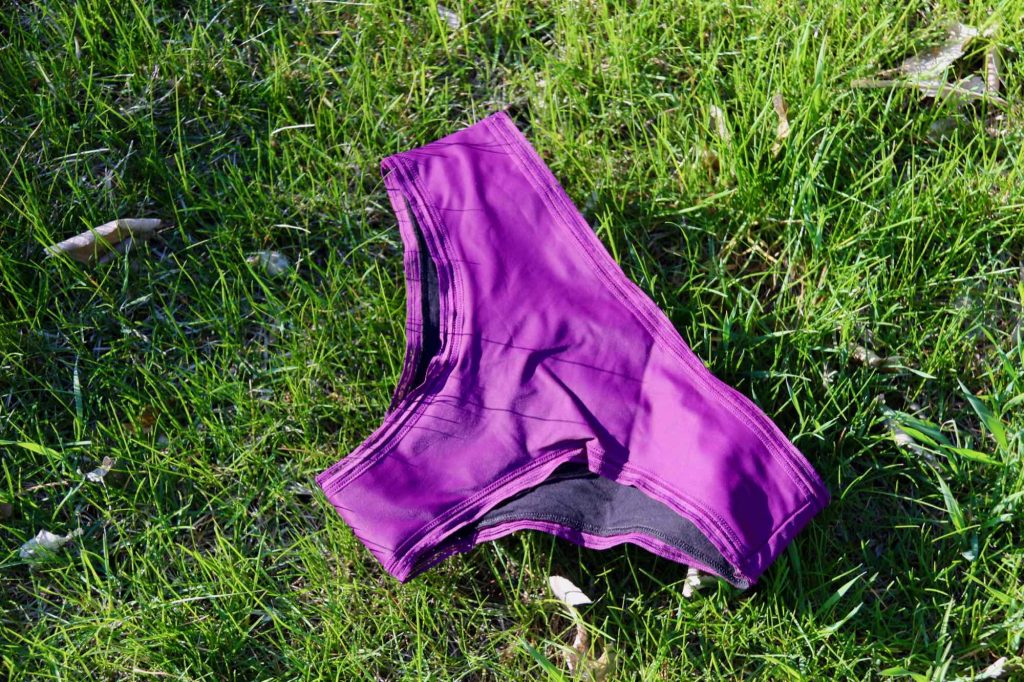 Thinx’s Cheeky underwear style, in the bright purple Fig color, lying flat in bright sunlight on a grassy yard. ©KettiWilhelm2021