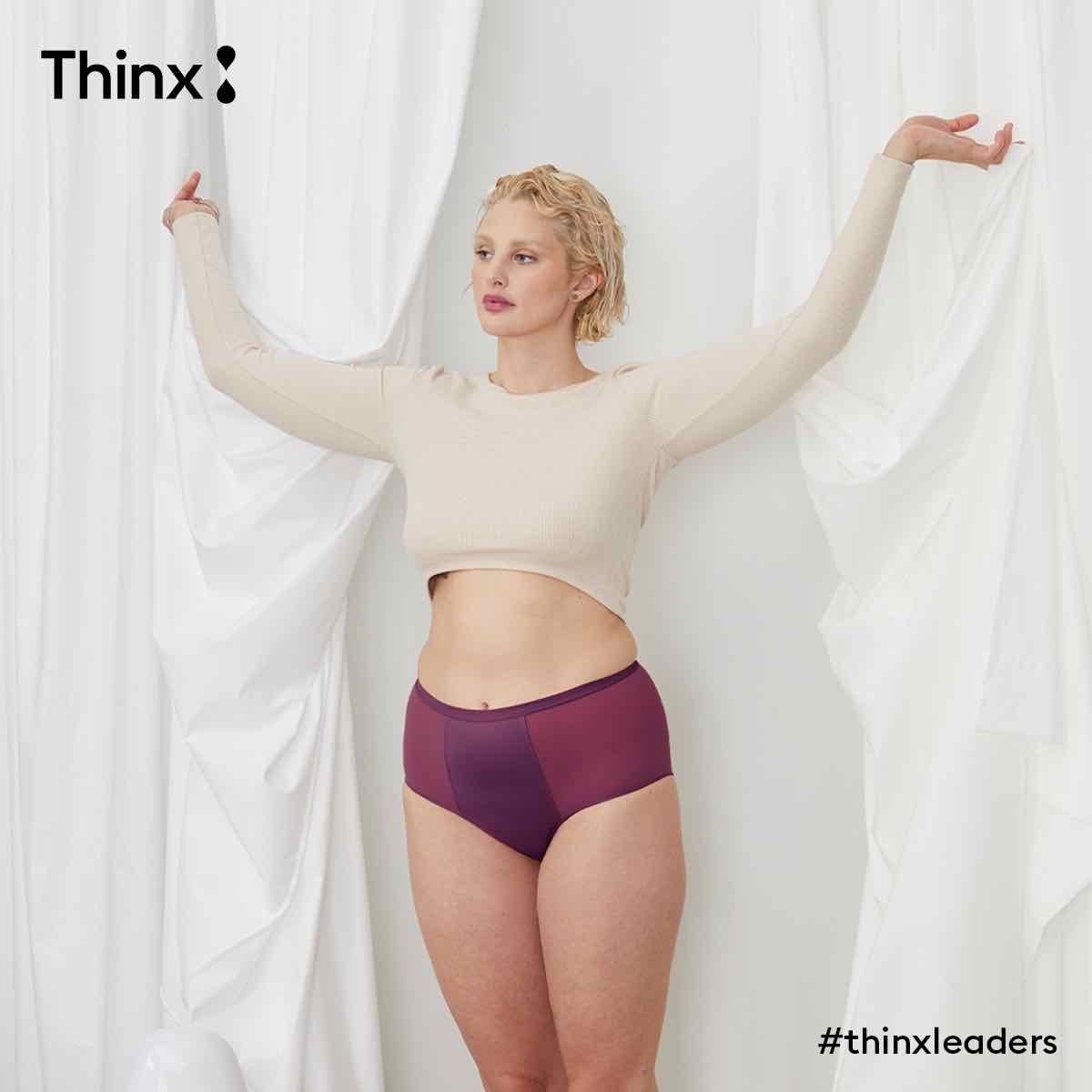 A model (a white woman with short blond hair) wearing a pair of Thinx hi-waist in the bright purple “fig” color, and a white sweater, looking away from the camera.
