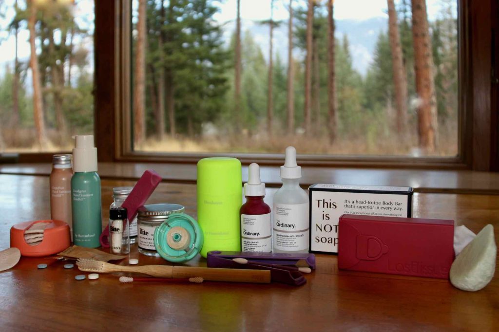 My zero-waste toiletry routine (including plastic-free deodorant, hand sanitizer, floss, and much more) sitting on a wooden table with a natural scene (trees and plants) in the background outside. ©KettiWilhelm2021
