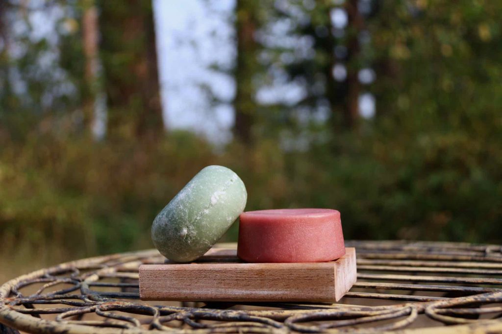 Two Earthling Co. bars – a green shampoo bar resting on top of a pink conditioner bar – in a natural setting, outside with trees in the background. ©KettiWilhelm2021