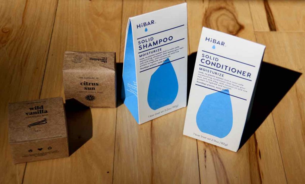 Displayed on a light-colored wooden surface are the compostable, recycled and recyclable paper packaging from Earthling Co. shampoo and conditioner bars (on the left) and HiBAR shampoo and conditioner bars (on the right).  ©KettiWilhelm2021