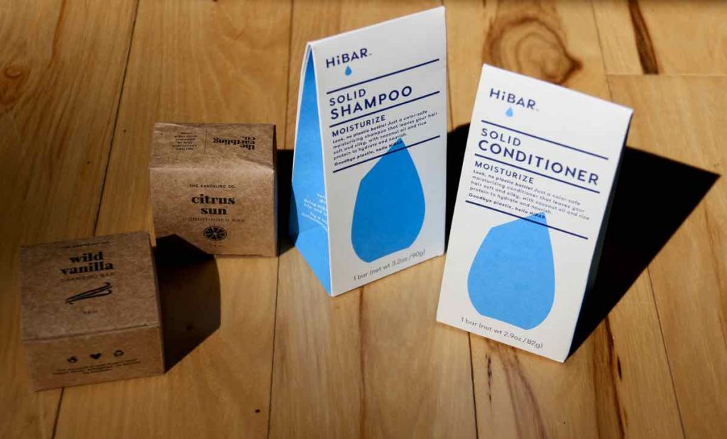 Displayed on a light-colored wooden surface are the compostable, recycled and recyclable paper packaging from Earthling Co. shampoo and conditioner bars (on the left) and HiBAR shampoo and conditioner bars (on the right). ©KettiWilhelm2021