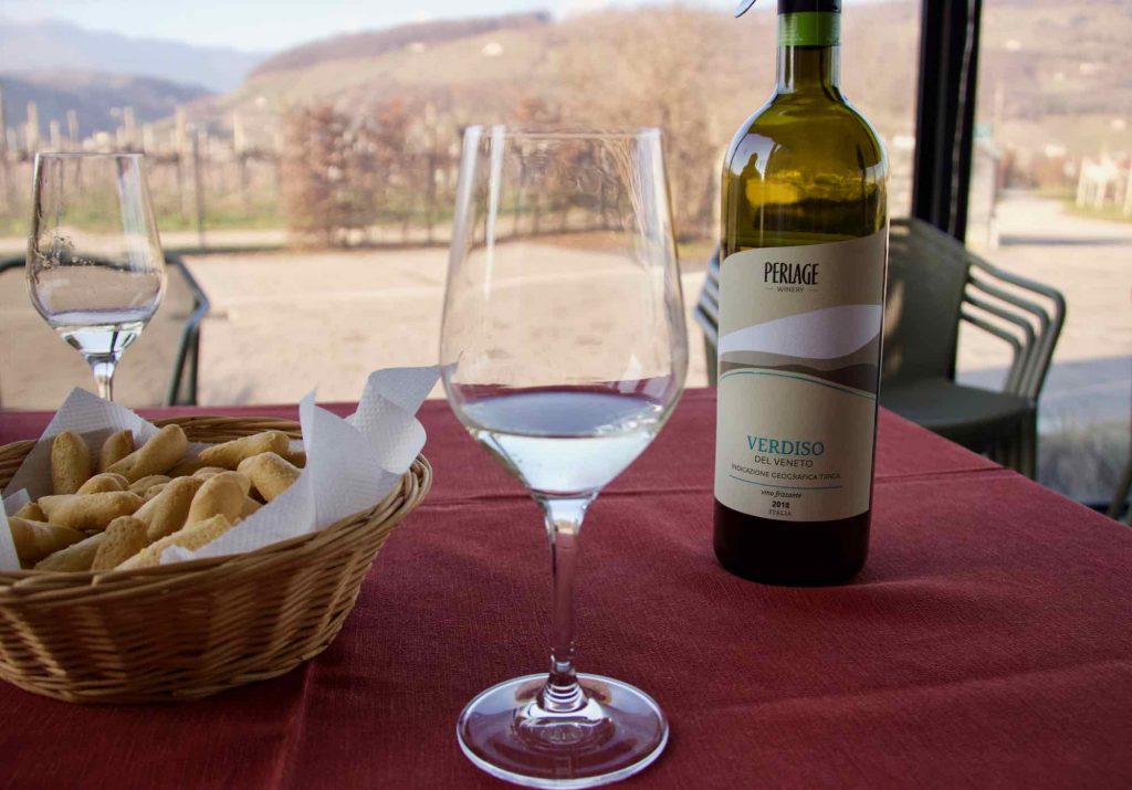 A glass of organic Prosecco at the Perlage Winery in Valdobbiadene, Italy – an easy choice to make a wine tasting trip more sustainable. A bottle of wine and basket of crackers are in the background. ©KettiWilhelm2021