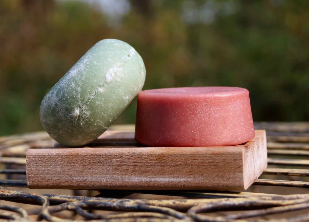 Green and pink shampoo and conditioner bars outdoors on a soap dish, with a green, natural background. Solid toiletries are an easy part of a sustainable travel routine. ©KettiWilhelm2021