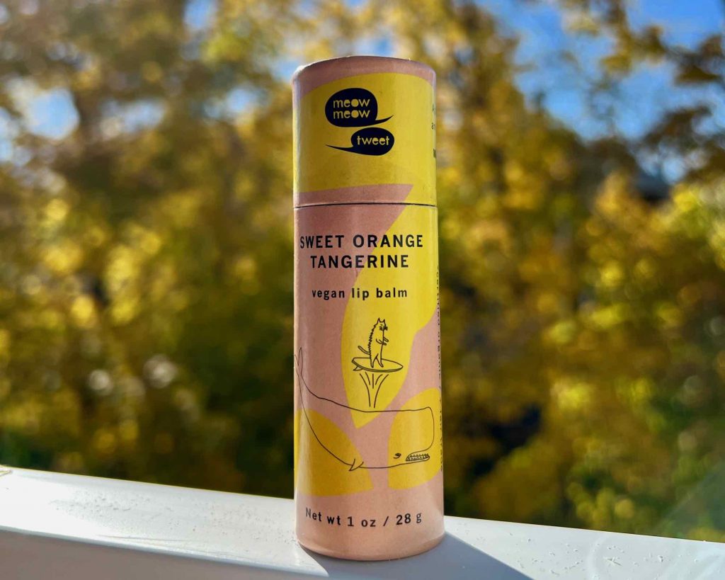 A colorful, pink and orange tube of Meow Meow Tweet’s vegan lip balm sits on a white window sill with bright blue and sky and fall foliage behind it. ©KettiWilhelm2021