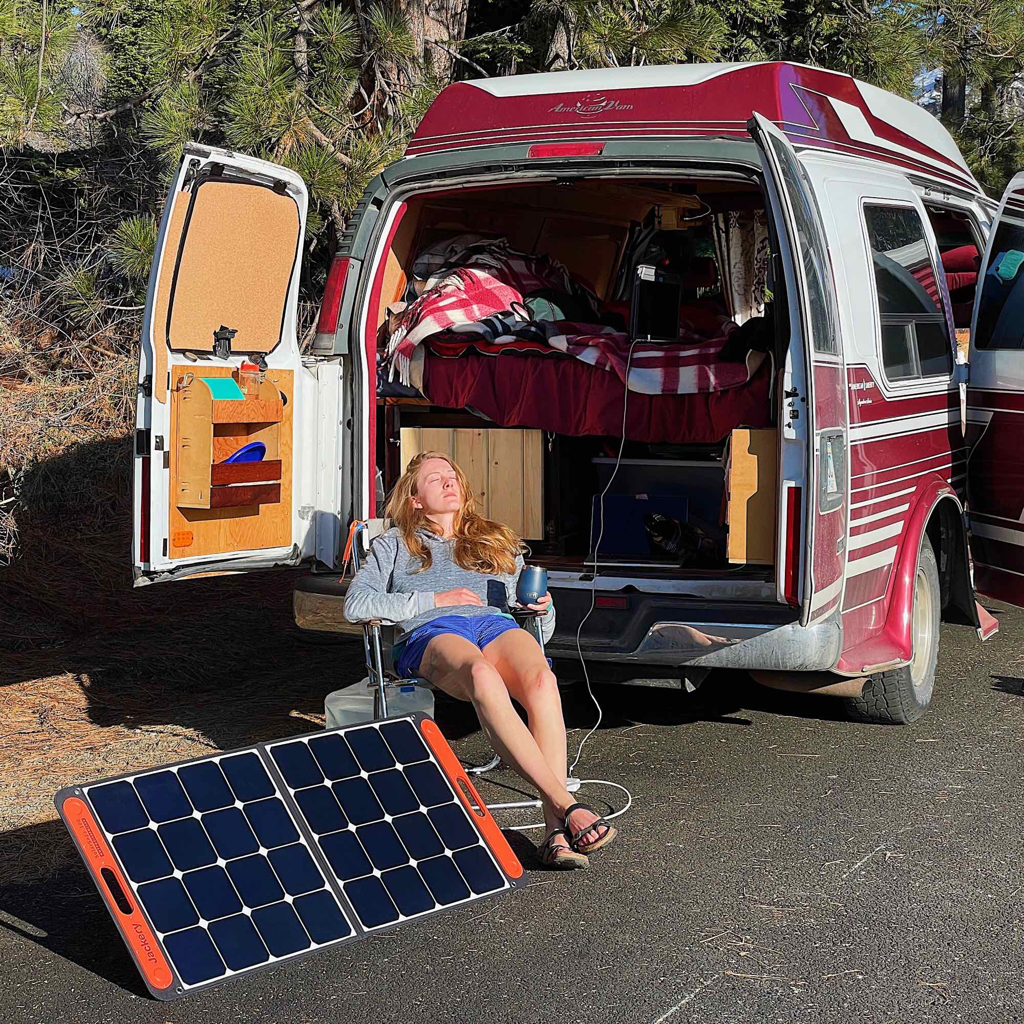 The author sitting on a fold chair in the sun, next to her Jackery solar panel, and converted van. ©KettiWilhelm2021