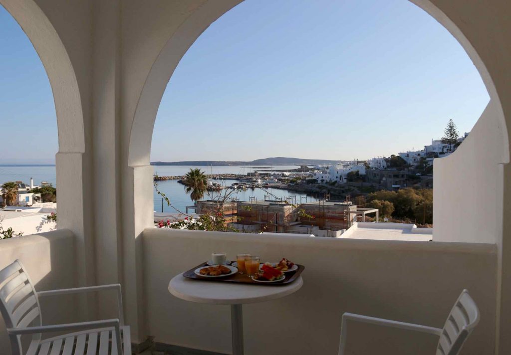 The view over the Mediterranean from the balcony at the Adonis Hotel, one of my favorite Clean Blue Paros hotels on the island, with a tray of Greek breakfast foods on the table. ©KettiWilhelm2021