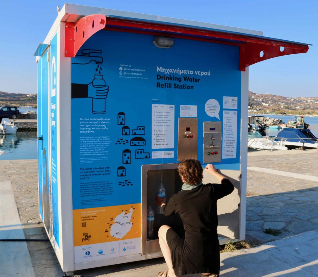 A woman refills a plastic water bottle from a blue filtered tap water refill station (about the size of car) on the island of Paros, where the NGO Clean Blue Paros recently redesigned the water stations to promote drinking tap water instead of bottled. ©KettiWilhelm2021