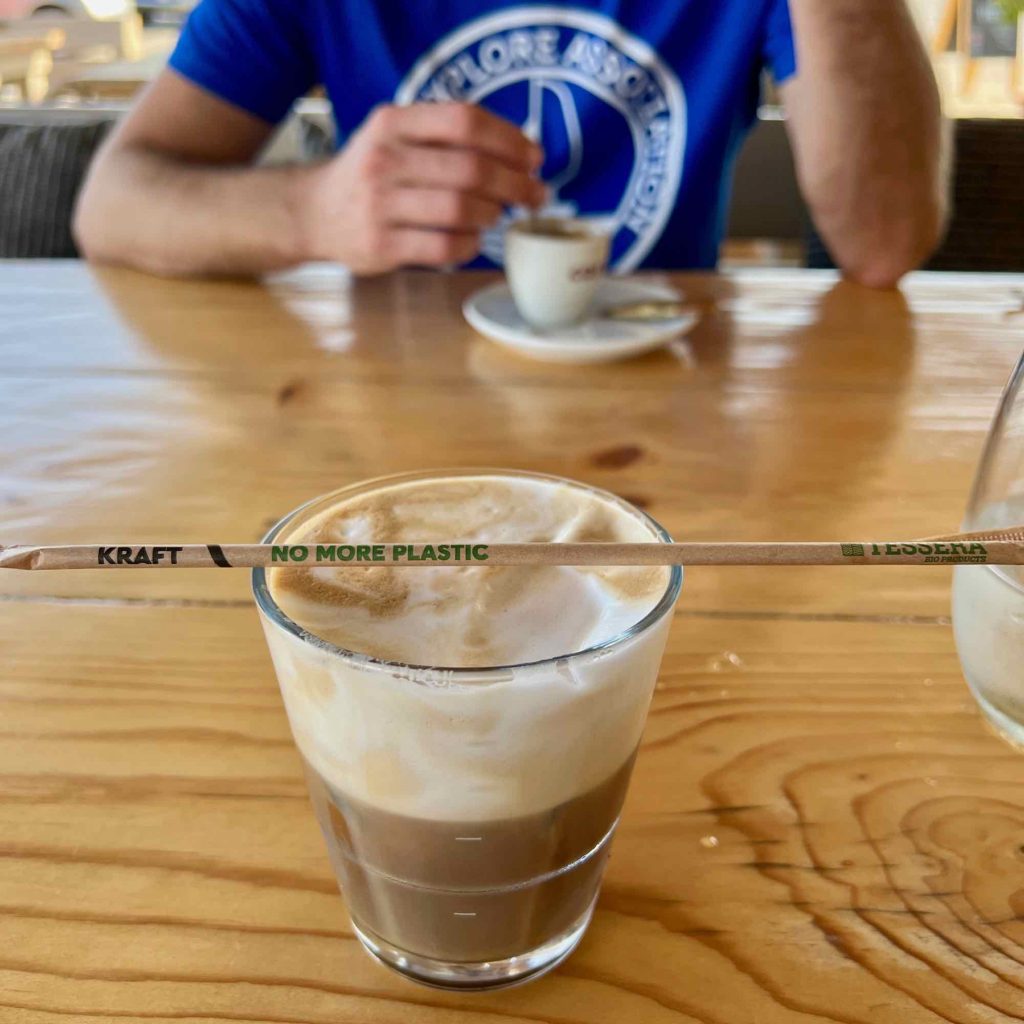 A classic Greek coffee cold drink, the “freddo cappuccino” (espresso with a thick layer of milk foam) in a glass with a paper straw on top that says “no more plastic.” ©KettiWIlhelm2021