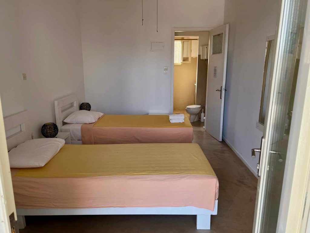 Inside the bedroom of my Standard Room at Okreblue Seaside Yoga Retreat on Paros: Plain white walls, two twin beds with peach-colored blankets, and no decoration. ©KettiWIlhelm2021