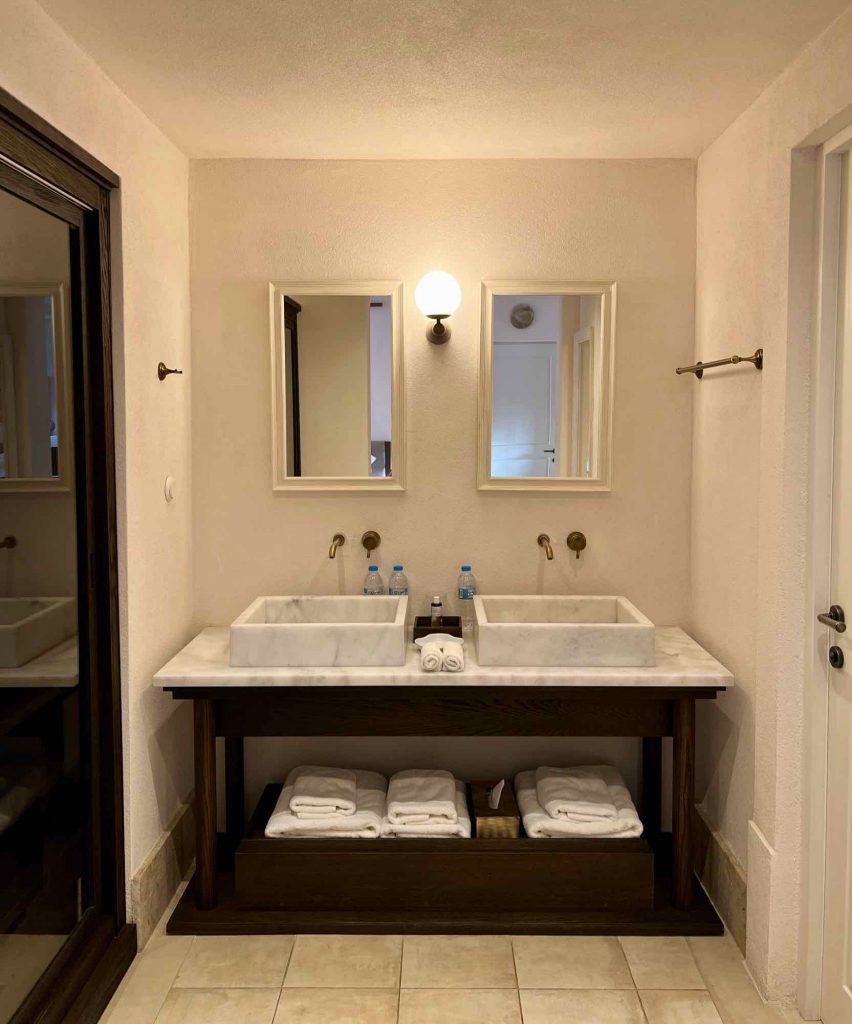 The bathroom in a Greek luxury hotel suite at Parilio, a Clean Blue Paros member business, with two white sinks and plastic water bottles. ©KettiWilhelm2021