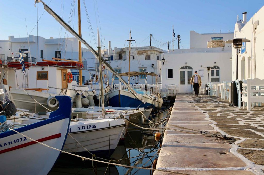 Sailboats docked at the port of Naousa, Paros, in the early morning, with an older, local man in the background wearing a mask. ©KettiWilhelm2021