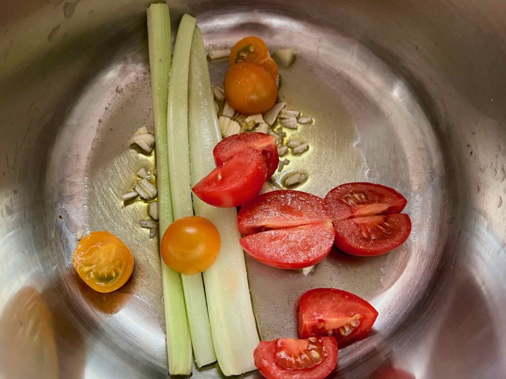Cherry tomatoes, a celery stick, minced garlic, salt, and olive oil lie at the bottom of a silver sauce pan. ©KettiWilhelm2022