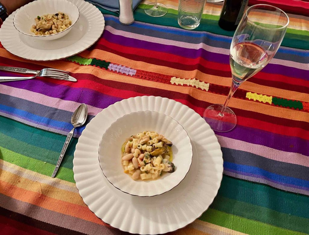 A plate of authentic Italian pasta with white beans and mussels on a white plate, on a brightly colored, striped tablecloth with a tall glass of Prosecco. ©KettiWilhelm2022
