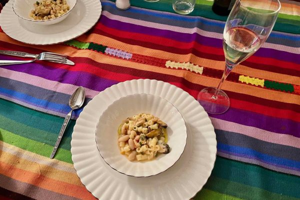 A plate of authentic Italian pasta with white beans and mussels on a white plate, on a brightly colored, striped tablecloth with a tall glass of Prosecco. ©KettiWilhelm2022