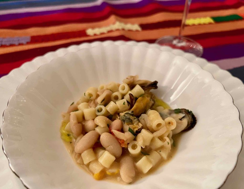 A plate of authentic Italian pasta with white beans and mussels on a white plate, on a brightly colored, striped tablecloth with a tall glass of Prosecco. ©KettiWilhelm2022