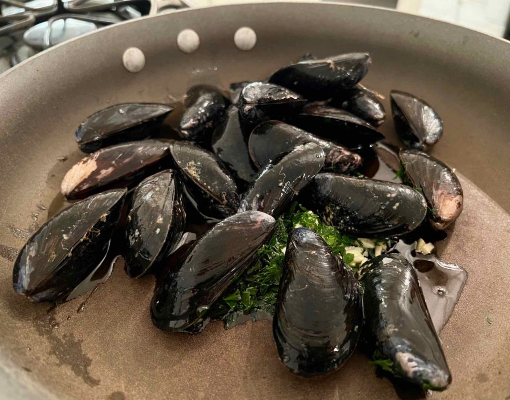 Close up of a pan with black-shelled mussels on top of minced garlic and parsley with olive oil. ©KettiWilhelm2022