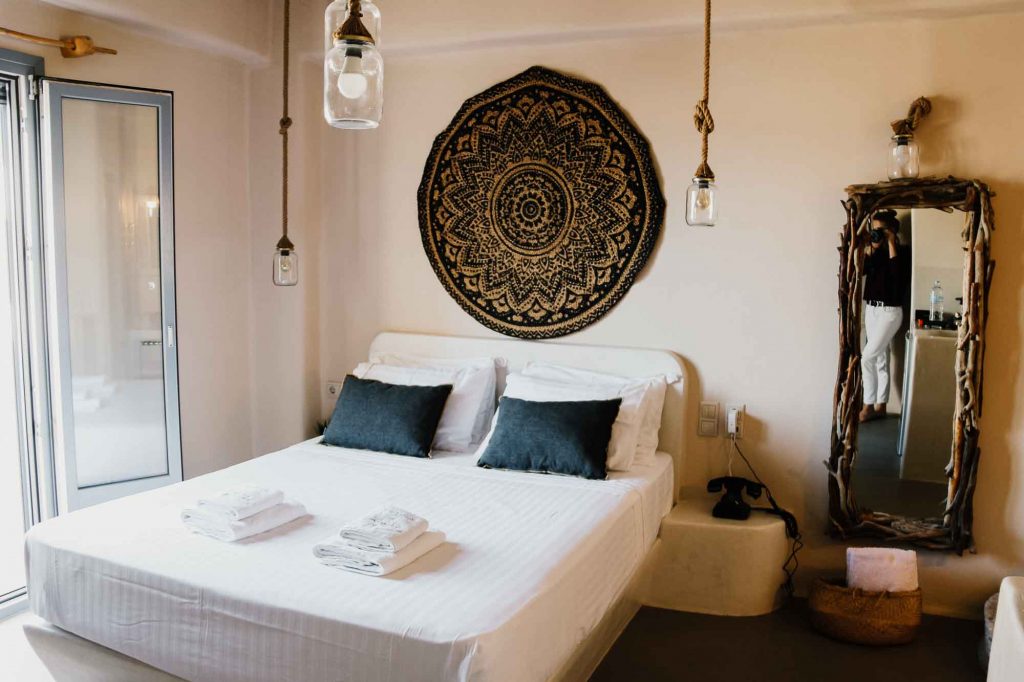 The bed in a room at the Ampelos Suites hotel in Naxos Town, on Naxos, Greece. A white comforter, black pillows, a decorative wicker art piece in black and tan, and a full-length mirror next to French doors that open onto a balcony. ©KettiWilhelm2022