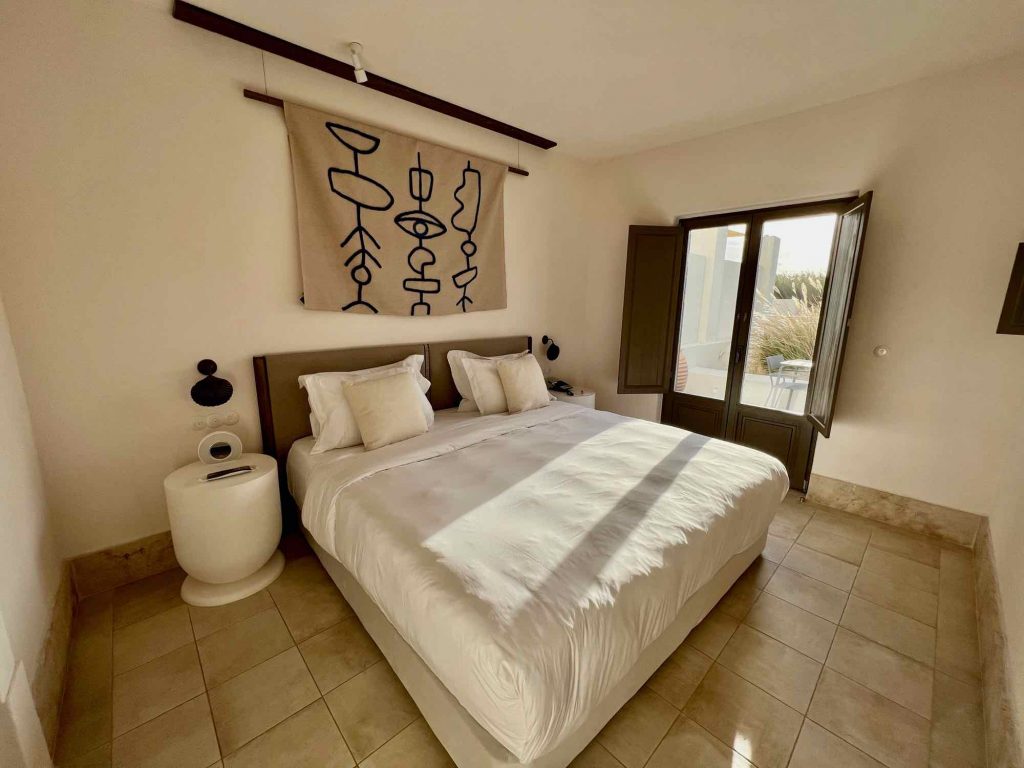 A bed with a white comforter in a neutral-colored hotel room at the luxurious Parilio Hotel on Paros, Greece. ©KettiWilhelm2022