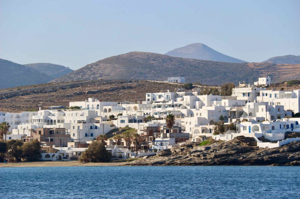 ALT The town of Naousa, on Paros, seen from a sailboat in the bay. Low, white buildings, and purple and blue mountains behind. ©KettiWilhelm2022