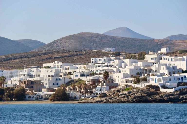 ALT The town of Naousa, on Paros, seen from a sailboat in the bay. Low, white buildings, and purple and blue mountains behind. ©KettiWilhelm2022
