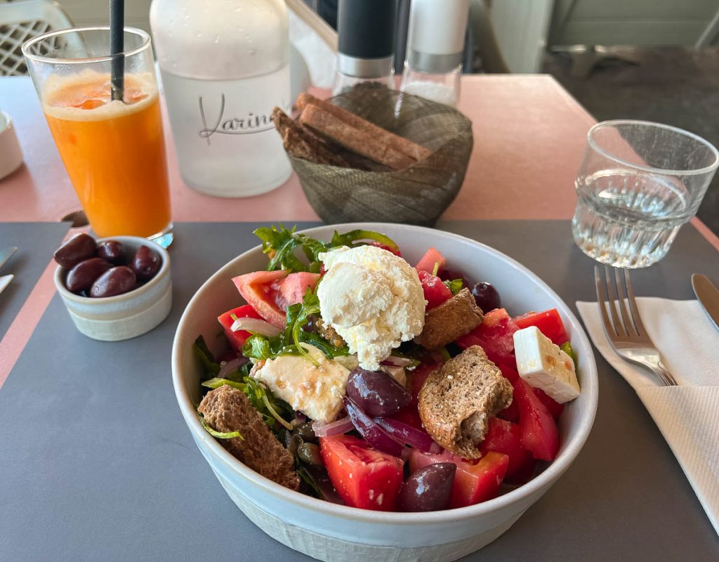 A Greek salad, a bowl of olives, and a glass of orange juice on a table with a refillable glass carafe of water at Café Karino in Naousa, Paros. ©KettiWilhelm2022