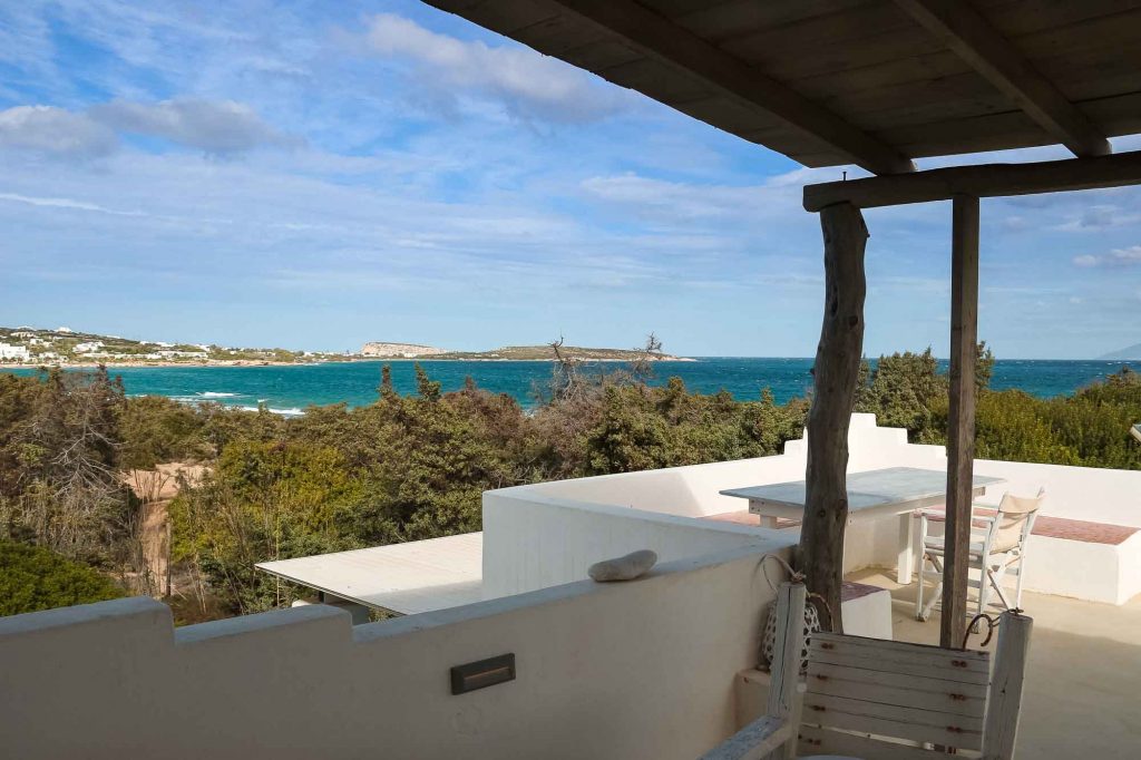 The sea view from the Tramountana Apartment at the Okreblue Yoga Retreat, one of the sustainable accommodation options in this travel guide to Paros. From under a wooden roof, a white patio looks out to the turquoise Mediterranean, with windblown trees in front. ©KettiWilhelm2022