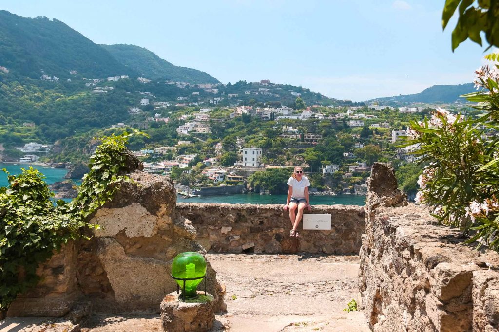 The author, perched on a castle wall in Ischia, with green mountains behind, working on her Italian habit of relaxation. ©KettiWilhelm2022