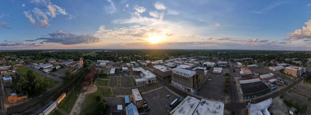 An aerial shot of downtown Clarksdale, Mississippi, at sunset. A freight train is on the left of the shot, and brick buildings, one to five stories tall, cover the rest of the town, while the sun hovers just above the horizon in the center. ©ChristineLozada2022