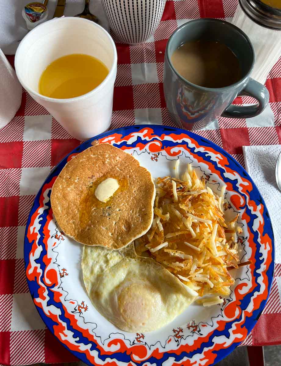 A cup of coffee in a ceramic cup, and a styrofoam glass of orange juice next to a bright, red, white and blue plate with pancakes, a fried egg, and hashbrowns, on a red and white checkered tablecloth at Our Grandma's House of Pancakes, Clarksdale, MS. ©KettiWilhelm2022