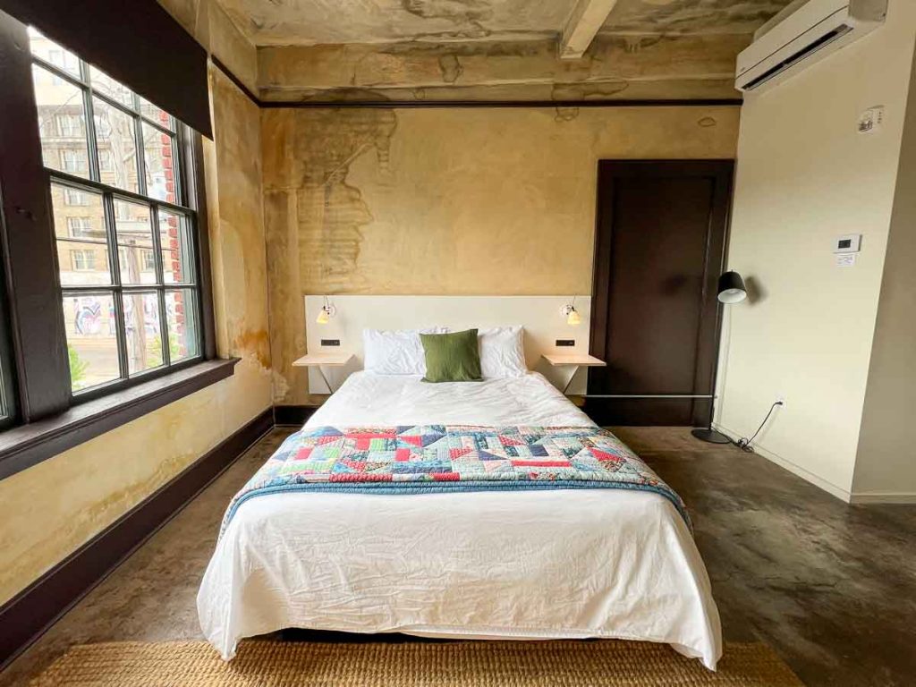 A bed at the locally-owned Travelers Hotel in Clarksdale, MS, with white covers, and a blue and yellow patchwork quilt folded at the foot, and naturally splotchy blue and yellow walls with lots of natural light from the large windows. ©KettiWilhelm2022
