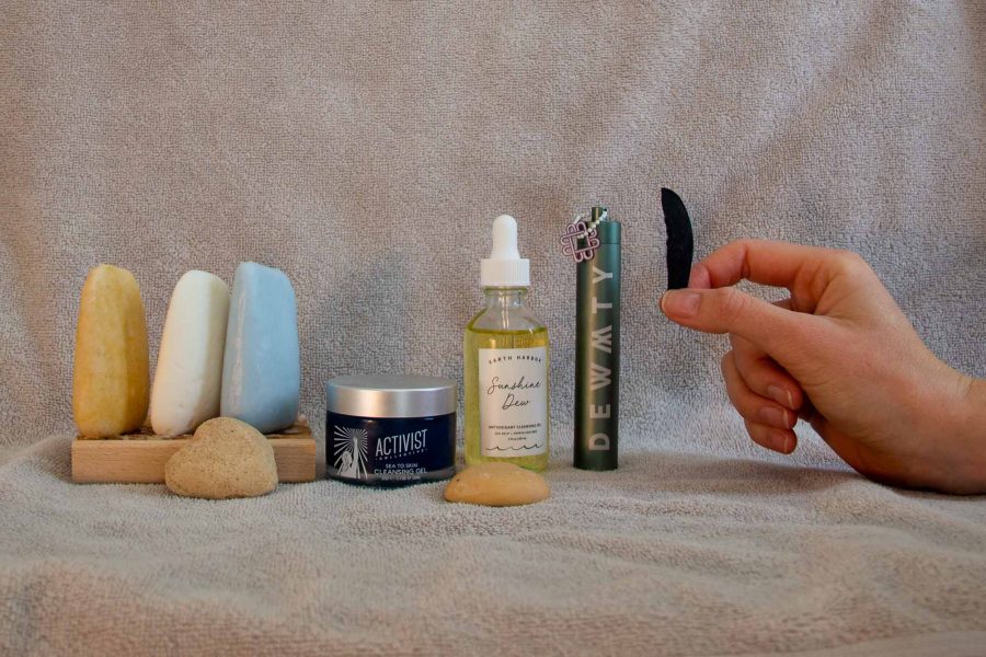 Plastic-free, zero-waste face wash options (bars, powders, and oils) from HiBAR, Earth Harbor, Activist Skincare, Dew Mighty, and Ethique sitting on a backdrop of a light gray towel. A white woman’s hand is shown holding up a small remaining piece of the long-lasting face wash bar from Earth Harbor. ©KettiWilhelm2022