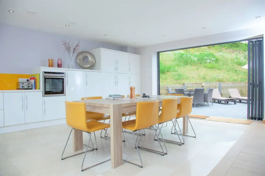 The bright kitchen inside this sustainable English design home in Devon – complete with bright yellow chairs around a light wooden table and a full wall of windows looking onto a green hillside.