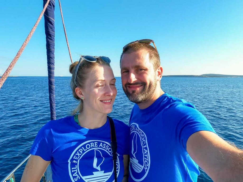 Selfie of the blogger Ketti Wilhelm, squinting into the camera with her husband Emanuele, during a trip with Sail and Explore, researching microplastics pollution in the Mediterranean. Both are wearing matching bright blue Sail and Explore t-shirts. ©KettiWilhelm2022