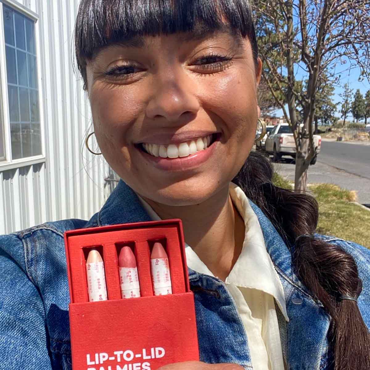 Ericka, founder of the sustainable small beauty company Axiology, holds her product in front of her while smiling for a selfie on the street.